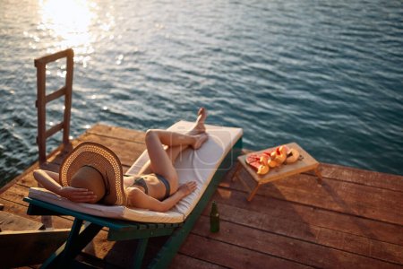Foto de Chica relajante en el muelle. Joven mujer atractiva en bikini en un muelle junto al agua durante el atardecer de verano - Imagen libre de derechos