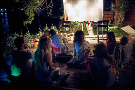 Téléchargez les photos : Un groupe de personnes assises sur le sol dans le bar sur la rive de la rivière et profitant du cinéma de nuit par une belle journée d'été. Nuit, été, bar, rivière - en image libre de droit