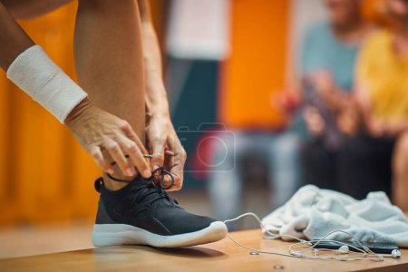 Foto de Mujer joven atándose el zapato en el vestuario del gimnasio, con auriculares y toalla en el banco, preparándose para el entrenamiento. Helath, estilo de vida, concepto activo. - Imagen libre de derechos
