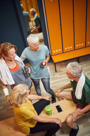 Photo for Vertical top shot of four people in gym locker room together enjoying talking before workout. Senior man and women with young woman trainer. - Royalty Free Image