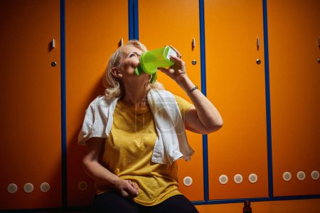 Foto de Hermosa mujer rubia senior sentada en el vestuario del gimnasio bebiendo agua después del entrenamiento. Salud, vida adulta, concepto de estilo de vida. - Imagen libre de derechos