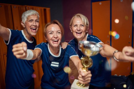 Photo for Team of two senior and one young women in dressing room, holding golden cup and feeling victorious. Team, togetherness, vicotry concept. - Royalty Free Image