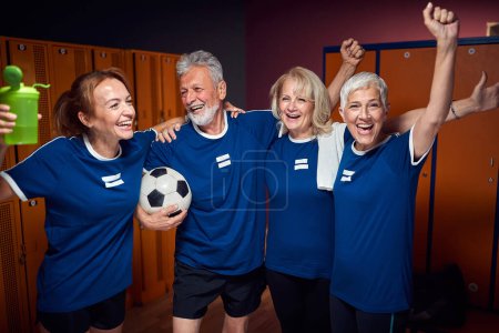 Photo for Football players in locker room celebrating together, senior man and woman with young trainer feeling joyful. Active lifestyle, togetherness, senior life concept. - Royalty Free Image