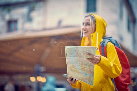 Foto de Mujer alegre con mapa en la lluvia mirando feliz. Camina por la ciudad con impermeable. Lluvia de verano. Viajes, concepto de estilo. - Imagen libre de derechos