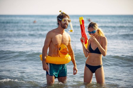 Foto de Loco joven con pato de goma y mujer sonriente con pistola de agua divirtiéndose en la playa - Imagen libre de derechos