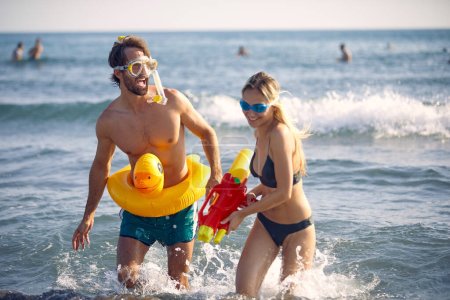 Cheerful young couple with watergun on the beach. Man with rubber duck pool float. Summer vacation concept.