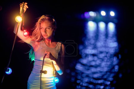 Téléchargez les photos : Jolie jeune femme blonde en robe d'été tenant des lampes aux guirlandes colorées la nuit. Festive, portrait, concept beauté. - en image libre de droit