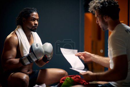 Photo for Muscular and strong boxing player in sweats after workout, resting on bench face to face with his coach, getting consultation. Sports, lifestyle concept. - Royalty Free Image
