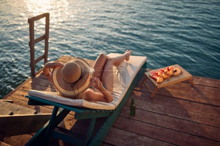 Foto de Mujer joven disfrutando tomando el sol en un embarcadero de madera junto al agua. Posando y sosteniendo su gran sombrero de paja de moda. Vacaciones, concepto de estilo. - Imagen libre de derechos