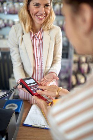 Foto de Mujer joven que paga con tarjeta de crédito, ingresando su código pin, encantadora persona de ventas sonriente sosteniendo lector de tarjetas de crédito para el cliente. Compras, concepto de estilo. - Imagen libre de derechos