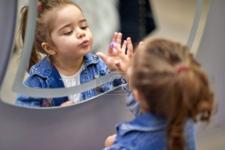 Foto de Adorable niña haciendo caras y mirándose en el espejo, tocando un botón en un espejo, espejo luminoso. Concepto de estilo de vida. - Imagen libre de derechos