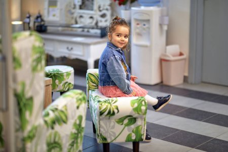 Photo for Adorable toddler girl sitting on modern upholstered chair in a furniture shop, looking around. Shopping, lifestyle concept. - Royalty Free Image