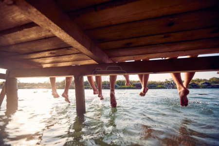 Foto de Cuatro personas con las piernas colgando al agua. Amigos en el agua de vacaciones. Diversión, unión, diversión, concepto de estilo de vida. - Imagen libre de derechos