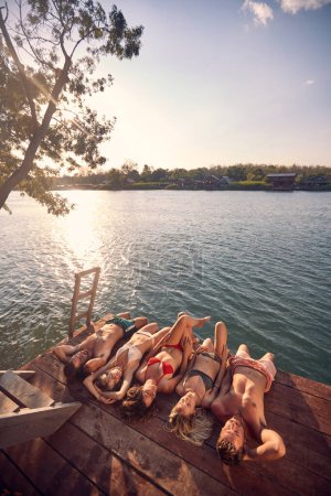 Foto de Cuatro amigos en traje de baño, acostados en embarcadero de madera por tomar el sol en el agua. Joven hombre y mujer de vacaciones. Vacaciones, unión, concepto de estilo de vida. - Imagen libre de derechos