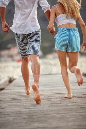 Téléchargez les photos : Un jeune couple court sur le quai au bord de la mer en se tenant la main par une belle journée. Amour, relation, vacances, mer - en image libre de droit