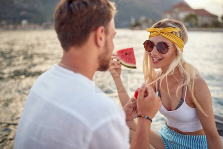 Photo for A young couple is enjoying a juicy watermelon while sitting on the dock on the seaside on a beautiful day. Love, relationship, holiday, sea - Royalty Free Image