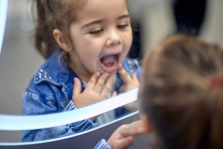Foto de Linda niñita mirando sus dientes en el espejo. Adorable niño pequeño haciendo caras y divirtiéndose en el espejo. Concepto de estilo de vida. - Imagen libre de derechos