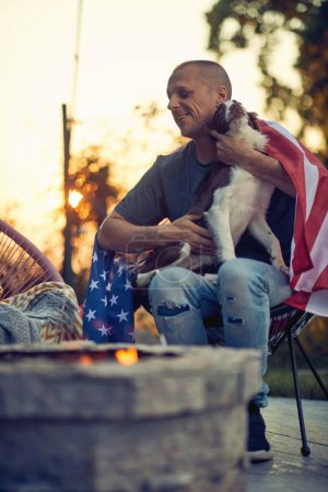 Foto de Alegre hombre de mediana edad con bandera de EE.UU. disfrutando de la noche con su perro cachorro, frontera collie al aire libre, sentado junto a una chimenea. Día de la Independencia. - Imagen libre de derechos