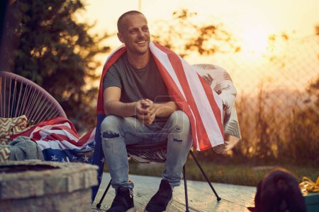 Photo for Wide horizontal shot of joyful young man with the flag of USA on his shoulders, relaxing in a cozy chair, outdoors in nature. Independence Day concept. - Royalty Free Image