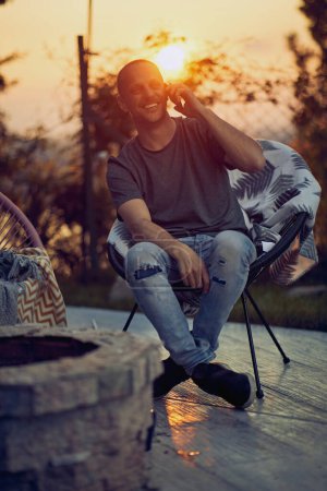 Téléchargez les photos : Joyeux jeune homme assis à l'extérieur dans une chaise confortable sur un patio, eith coucher de soleil en arrière-plan, parler au téléphone, se sentir joyeux. Style de vie, concept de relaxation. - en image libre de droit