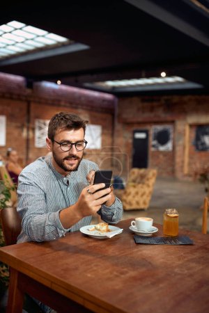 Photo for Young man with glasses, in cafe, looking at his phone, scrolling. Business, student, coffee break concept. Modern interior background. - Royalty Free Image