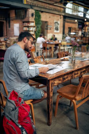 Foto de Joven barbudo en ropa casual con bolso de mochilero a su lado, sentado solo en la mesa leyendo el periódico. Pausa para café. - Imagen libre de derechos