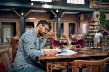Téléchargez les photos : Beau jeune homme barbu avec un verre assis seul dans un café lisant un journal. Pause café dans un café moderne. - en image libre de droit