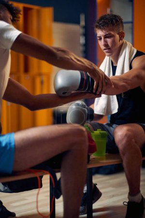 Photo for Young handsome boxing player getting ready for workout with his coach in dressing room, sitting on bench, putting on gloves. Sports, martial arts, lifestyle concept. - Royalty Free Image