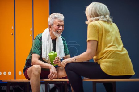 Photo for Sporty mature man and woman in gym locker room sitting facing each other and having a joyful conversation. Senior life, health, lifestyle concept. - Royalty Free Image