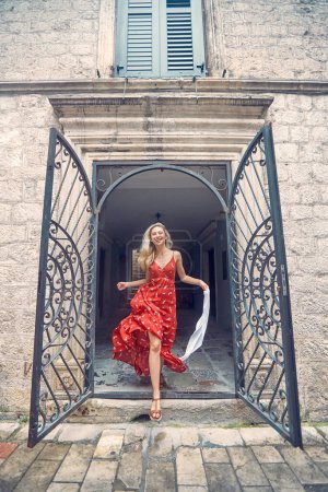 Foto de Rubia alegre saltando en vestido sensual rojo, sintiéndose feliz. Antiguo edificio. Vacaciones de verano, moda, estilo de vida concepto. - Imagen libre de derechos