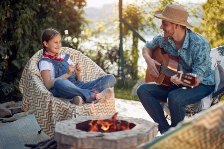 Seated by a crackling fire, the father strums a classical guitar, filling the air with beautiful melodies. His young daughter holds a marshmallow and smiles.