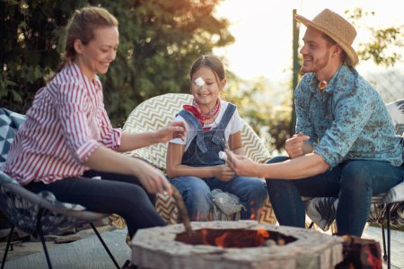 Photo for Gathered around the warm glow of the fire, a loving mother, father, and their daughter engage in the enjoyable activity of roasting marshmallows. - Royalty Free Image