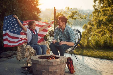 Foto de Celebración familiar del Día de la Independencia. El padre y su hija pequeña se sientan juntos junto al fuego, disfrutando del proceso de asar malvaviscos. La hija orgullosamente exhibe una bandera americana desplegada. - Imagen libre de derechos