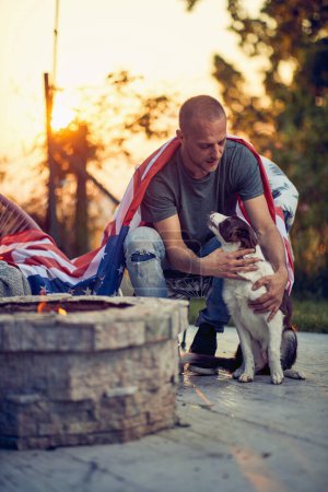 Téléchargez les photos : Jeune homme caressant chien chiot collie frontière, profiter du temps à l'extérieur, avec drapeau des États-Unis sur le dos, assis près d'une cheminée dans une chaise confortable. Jour de l'indépendance. - en image libre de droit