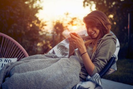 Photo for Cheerful young woman using smartphone, laying outdoors in a cozy chair with blanket on, enjoying evening. Sunset background. - Royalty Free Image