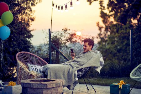 Téléchargez les photos : Joyeux jeune homme assis sur une chaise confortable avec une couverture, en utilisant un smartphone, détente en plein air dans un chalet. Coucher de soleil et fond nature. - en image libre de droit