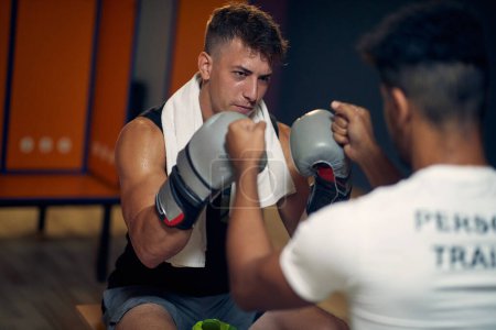 Photo for Determined and focused young sweaty boxing player with his coach in dressing room sitting on bench, feeling powerful. Sports, lifestyle, martial arts concept. - Royalty Free Image