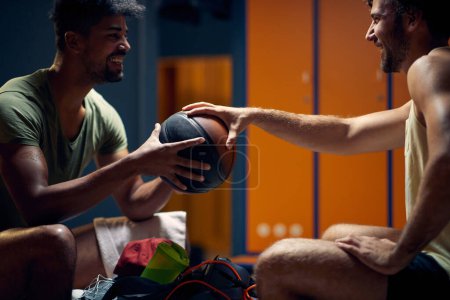 Téléchargez les photos : Joyeux jeunes hommes assis face à face dans les vestiaires de la salle de gym, tenant le ballon de basket et parlant. Préparation à l'entraînement. - en image libre de droit