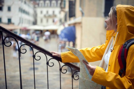 Foto de Viajar en la ciudad bajo la lluvia. Mujer joven con impermeable amarillo escondido de la lluvia, mirando hacia fuera y sonriendo. - Imagen libre de derechos