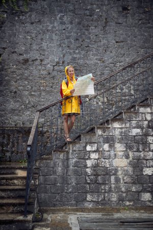 Téléchargez les photos : Une jeune touriste utilise une carte tout en marchant dans la vieille ville pendant les vacances un jour de pluie sur le bord de la mer. Vacances, tourisme, bord de mer - en image libre de droit