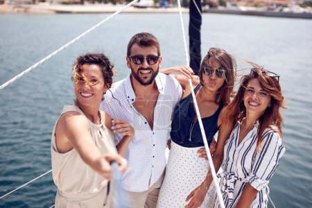 Téléchargez les photos : Un groupe de jeunes beaux modèles prend un selfie sur le pont du yacht tout en traversant le quai par une belle journée d'été sur le bord de la mer. Été, mer, vacances, amitié - en image libre de droit