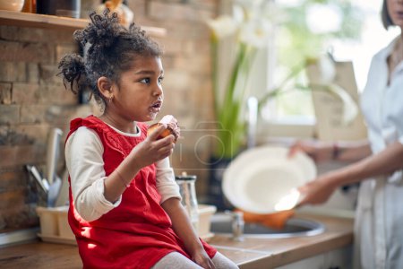 Téléchargez les photos : Petite fille assise dans la cuisine, se délectant d'un muffin appétissant, tandis que sa mère se tient à côté d'elle, laver la vaisselle avec diligence. - en image libre de droit