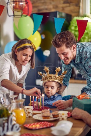 Téléchargez les photos : La petite fille fête joyeusement son anniversaire entourée de ses parents adorés.Sa mère allume soigneusement les bougies sur le gâteau joliment décoré, ajoutant une touche de magie au moment spécial. - en image libre de droit