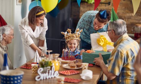 La niña está celebrando alegremente su cumpleaños. Rodeada de sus amorosos padres, abuelos y el pastel de cumpleaños adornado con velas, la escena está llena de felicidad y anticipación.