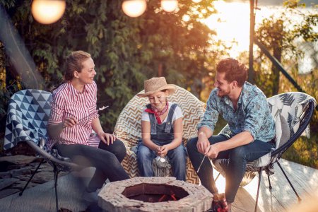 Photo for Family of three sitting and enjoying roasting marshmallows together by the fireplace, mother, father and daughter together. Lifestyle, vacation, togetherness concept. - Royalty Free Image