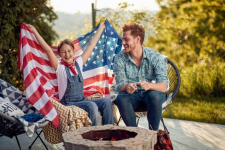 Photo for Joyful young girl holding USA flag, sitting outdoors with her father enjoying sunny summer day together. Independence day concept. - Royalty Free Image