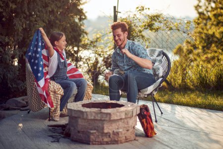 Foto de Hombre y niña sentados al aire libre junto a una chimenea asando malvaviscos, niña sosteniendo la bandera de EE.UU., celebrando juntos el día de la Independencia. Vacaciones, familia, concepto de unión. - Imagen libre de derechos