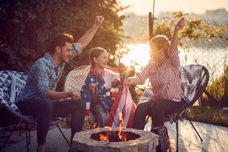 Photo for Beautiful and joyful family of three sitting outdoors by a fireplace, roasting smores, cheering, enjoying sunny summer day together. Fourth of July, Independence Day concept. - Royalty Free Image