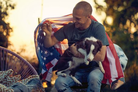 Photo for Middle age man with USA flag on his back cuddling with puppy border collie, outdoors in a cozy chair, at sunset. 4th of July, Independence Day - Royalty Free Image