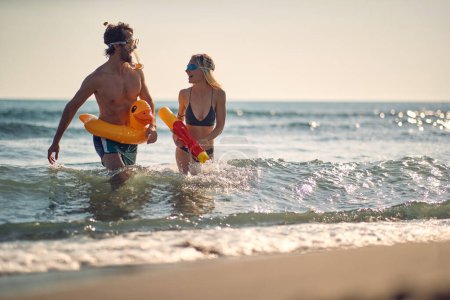 Playful young attractive couple on beach. Woman with water gun, man with rubber duck pool float. Holiday, summertime, lifestyle concept.
